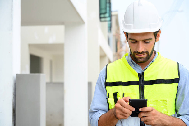 portrait of young engineer in vest with white helmet standing on construction site, smiling and holding smartphone for worker, internet, social media - Фото, изображение