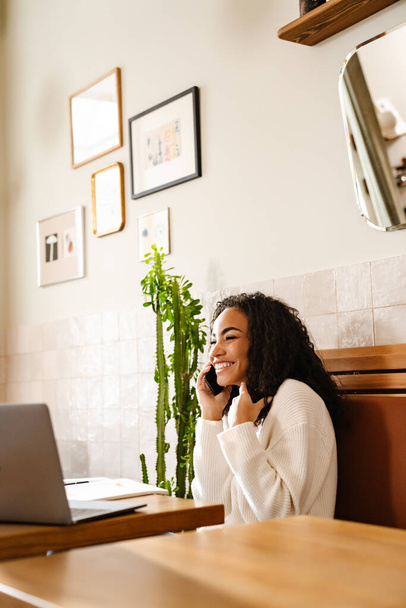 若い黒人女性が携帯電話で話をしながら、カフェでラップトップで屋内で働く - 写真・画像