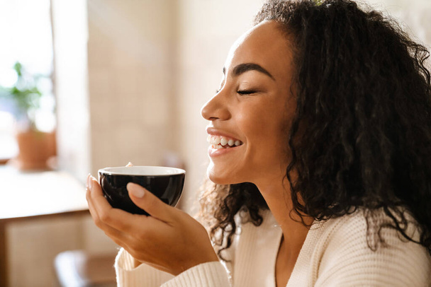 Young black woman laughing and drinking coffee while sitting in cafe indoors - Φωτογραφία, εικόνα