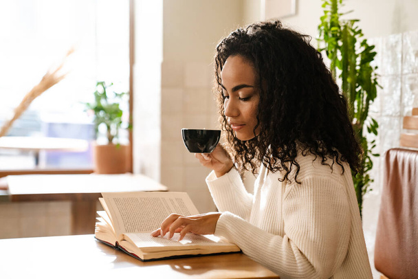 Young black woman drinking coffee and reading book in cafe indoors - Fotoğraf, Görsel