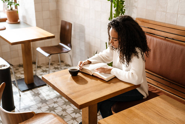 Young black woman drinking coffee and reading book in cafe indoors - Фото, изображение