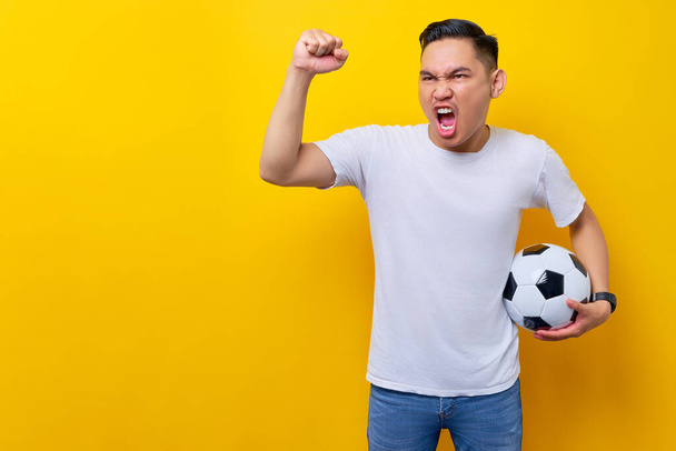 fanoušci nadšeni podporou fotbalového sportovního týmu. Asijský muž 20s nosí bílé tričko drží fotbalový míč a sledování živého proudu v televizi s dělat vítězný gesto izolované na žlutém pozadí - Fotografie, Obrázek