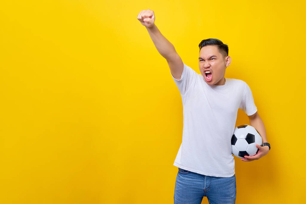 fanit innoissaan tukemaan jalkapallojoukkuetta. Aasialainen mies 20s pukeutuu valkoinen t-paita tilalla jalkapallo pallo ja katsella live stream televisiosta tekee voittaja ele eristetty keltaisella pohjalla - Valokuva, kuva