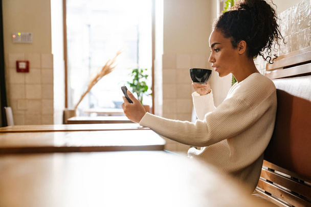 喫茶店内でコーヒーを飲みながら携帯電話を使う若い黒人女性 - 写真・画像