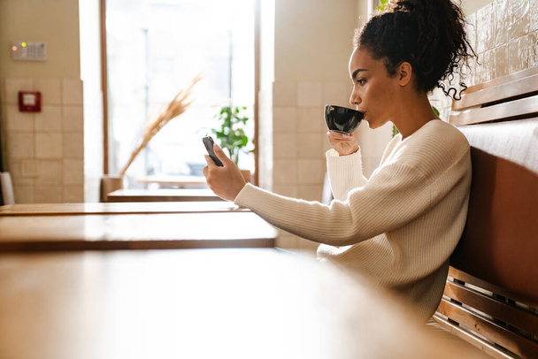 Young black woman using mobile phone while drinking coffee in cafe indoors - Foto, Imagem