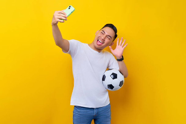 ενθουσιασμένοι νεαρός ασιάτης οπαδός του ποδοσφαίρου σε ένα λευκό t-shirt μεταφέρουν μια μπάλα ποδοσφαίρου και να κάνει μια selfie πλάνο σε ένα κινητό τηλέφωνο απομονώνονται σε κίτρινο φόντο. Οι άνθρωποι αθλητισμός αναψυχής έννοια τρόπο ζωής - Φωτογραφία, εικόνα