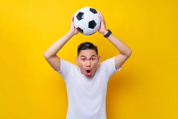 Cheerful young Asian man footbal fan in white t-shirt cheer up support favorite team catching soccer ball gesture isolated on yellow background. People sport leisure lifestyle concept - Valokuva, kuva