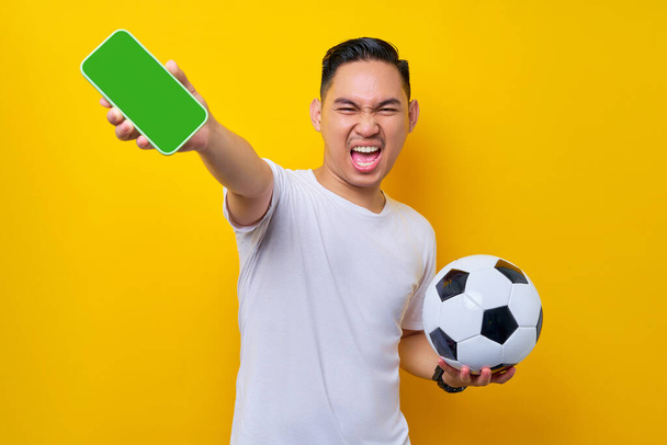 podekscytowany młody Azjata kibic w białym t-shircie trzymając piłkę nożną i pokazując pusty ekran telefon komórkowy odizolowany na żółtym tle. Ludzie sport rekreacyjny styl życia koncepcja - Zdjęcie, obraz