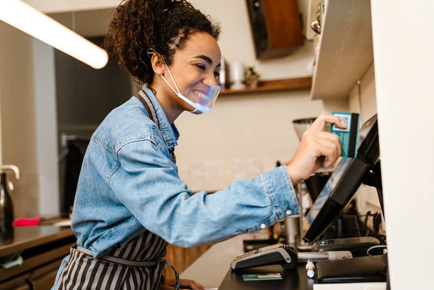 Black barista woman wearing face mask working with cash register in cafe indoors - Zdjęcie, obraz