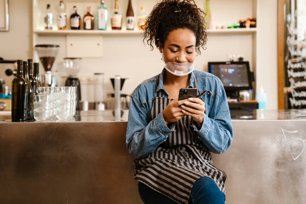 Young black waitress in face mask using mobile phone while working at cafe indoors - 写真・画像