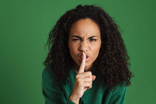 Young black woman frowning and making silence gesture isolated over green background - 写真・画像