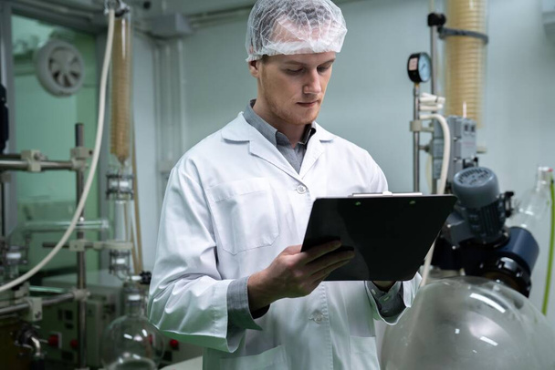 Apothecary scientist using a clipboard and pen to record information from a CBD oil extractor and a scientific machine used to create medicinal cannabis products. - Photo, Image
