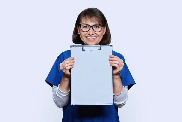 Woman doctor pointing finger at blank paper on clipboard, white studio background. Friendly middle aged nurse in blue uniform, clipboard space for personal message advice information advertising text - Fotó, kép
