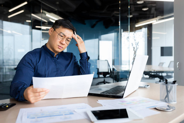 Desperate businessman looks and reads financial reports and documents, Asian boss works with documents inside office, confused and depressed. - 写真・画像
