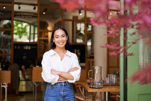 Πορτρέτο της νεαρής επιχειρηματία στο δικό της καφέ, διευθυντής στέκεται κοντά στην είσοδο και σας προσκαλεί, ποζάροντας σε λευκό απλό πουκάμισο και τζιν. - Φωτογραφία, εικόνα