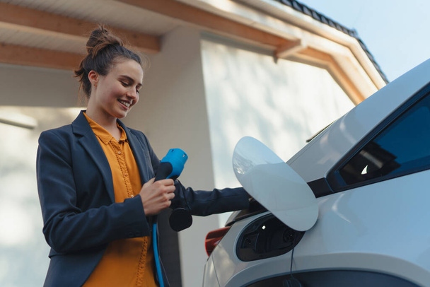 Young woman holding power supply cable from her car, prepared for charging it in home, sustainable and economic transportation concept. - Fotografie, Obrázek