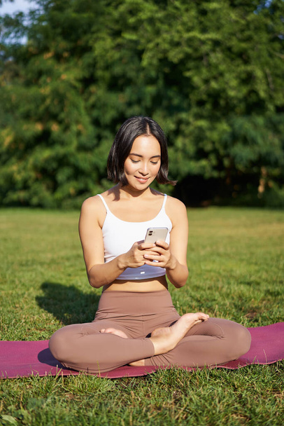 Portrait of korean girl meditating in park, follow yoga video lesson on smartphone app, sitting on fresh air and relaxing. - Photo, Image