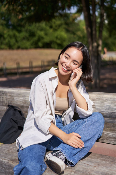 Χαμογελαστή Ασιάτισσα που χαλαρώνει στο πάρκο, μιλάει σε smartphone, έχει κινητό ενώ ξεκουράζεται στο ύπαιθρο. - Φωτογραφία, εικόνα