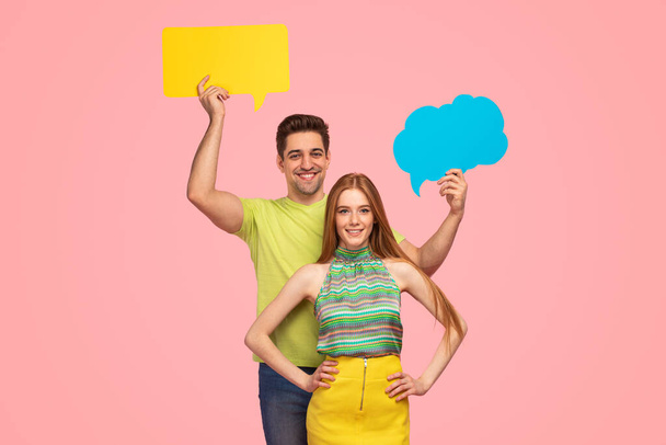 Happy man smiling and demonstrating bright speech bubbles while standing behind girlfriend in stylish colorful clothes against pink background - Фото, изображение