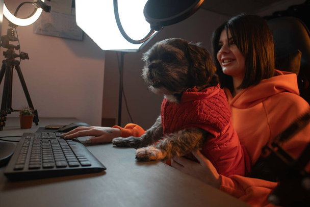 gracz dziewczyna i jej szczeniak są streaming na żywo w Internecie, ma szczeniaka na kolanach, ma na sobie pomarańczową bluzę i jest w pokoju gier strumieniowo z pierścieniem świetlnym. - Zdjęcie, obraz