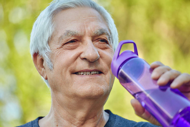 Close up face attractive mature man holds plastic reusable bottle drinking still mineral water during morning work out or stroll in summer park, caring about health, relieving thirst - Foto, Imagem