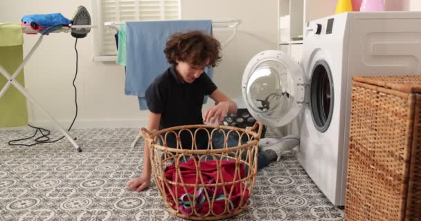 A smiling teen boy in jeans and a shirt sits in front of a home washing machine. He loads dirty clothes into the washing machine. A bright and airy laundry room. - Footage, Video