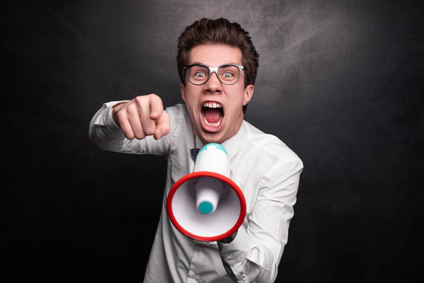 Angry male leader in smart casual clothes and glasses with megaphone pointing at camera and screaming against black background - Foto, immagini