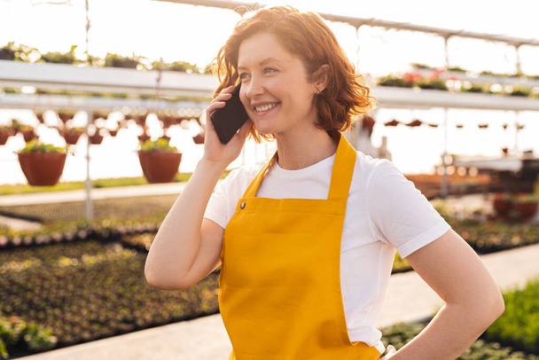 Happy woman in white t shirt and yellow apron smiling and answering phone call while working in hothouse at sunset - Foto, immagini