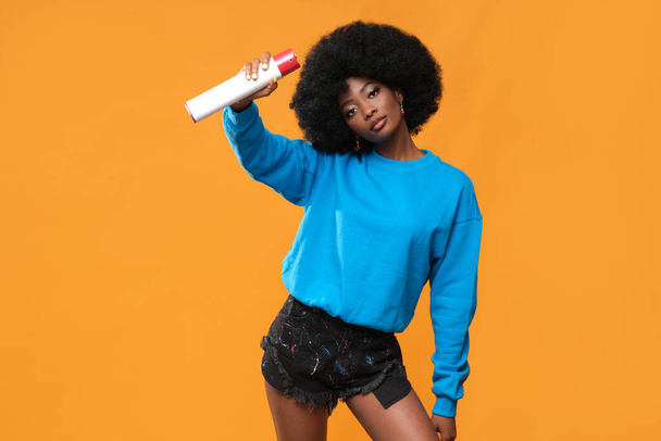 Black-skinned model with an afro hairstyle and spray paint in her hand on an orange background. - Φωτογραφία, εικόνα