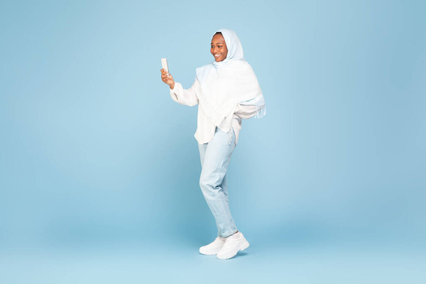 Happy african american woman in hijab using cellphone, standing on blue studio background, free space. Black lady chatting online, browsing social media, taking photo, full body length - Photo, Image