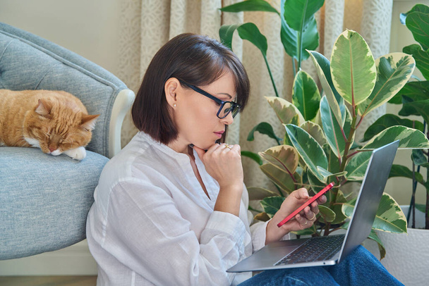 Middle aged woman sitting on floor using smartphone laptop. Female in home interior with sleeping pet ginger cat on armchair. Work at home, technology, leisure, freelancing, lifestyle, people, animal - Zdjęcie, obraz