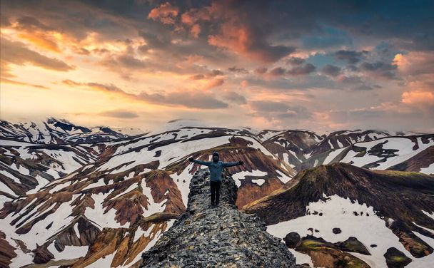 Erfolgreicher Wanderer, der mit erhobenen Armen auf dem Felsen mit vulkanischem Berg und Schnee vom Blanhjukur-Pfad steht, an einem Sommerabend in Landmannalaugar, Island - Foto, Bild