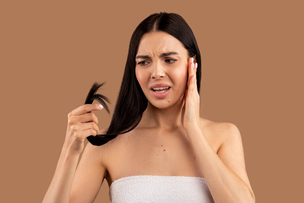 Anxious beautiful half-naked young brunette woman checking her long hair, holding split ends and touching her face, beige studio background, copy space. Hair care natural organic treatment concept - Photo, Image