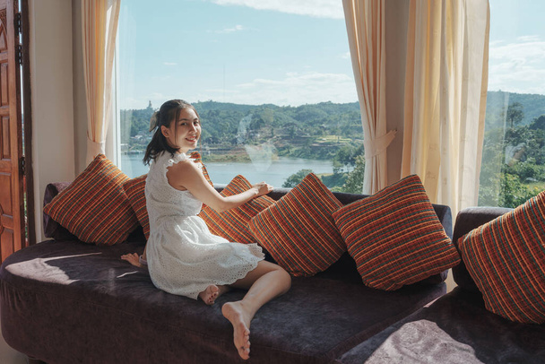 Cute asian woman relaxing and enjoying the tropical forest view on sofa at panorama room in bright day on summer vacation - Photo, Image