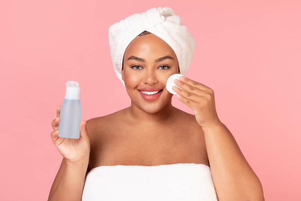 Retrato de una hermosa mujer obesa negra limpiando su piel con almohadilla de algodón, sosteniendo la botella con tóner y sonriendo a la cámara sobre el fondo rosa del estudio, espacio para copiar - Foto, imagen