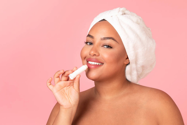 Chapped lips prevention. Beautiful black oversize woman using chapstick and smiling at camera, posing wrapped in towel and applying Llip balm over pink background, copy space - Foto, afbeelding