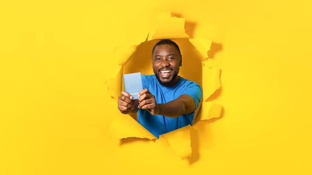 Happy black male traveler tourist holding passport and smiling at camera, posing in torn hole of yellow paper wall background, free copy space, panorama - Fotografie, Obrázek