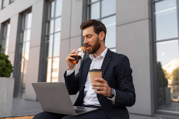 Ernsthafter junger europäischer Manager mit Bart im Anzug schaut sich Video auf Laptop mit Tasse Kaffee zum Mitnehmen an, isst Croissant, sitzt auf einer Bank in der Nähe eines Gebäudes in der Stadt. Mittagessen im Freien, Pause von der Arbeit, Geschäft - Foto, Bild