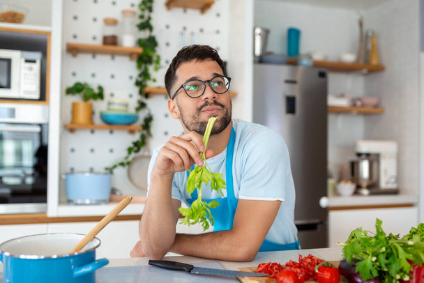 Handsome smiling young man leaning on kitchen counter with vegetables and looking away. Portrait of happy casual guy in apron leaning on steel counter in the kitchen with ingredients on it. - Foto, imagen