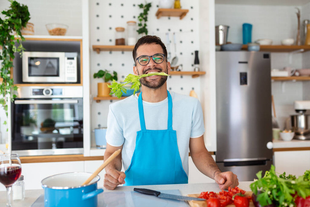 Счастливый красавец, готовый на кухне дома, с сельдереем во рту, позирующий для фото, концепция здорового образа жизни - Фото, изображение