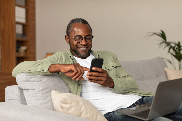 Рад среднего возраста африканский американец в очках читает сообщение по телефону, смотрит видео-серфинг в интерьере комнаты, копирует пространство. Приложение для социальных сетей, блога, поиска в интернете и нового устройства дома - Фото, изображение