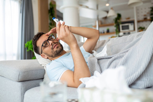 Man feeling sick lying in the bed and looking the thermometer. Sick man lying on sofa checking his temperature under a blanket at home in the living room - Foto, Bild