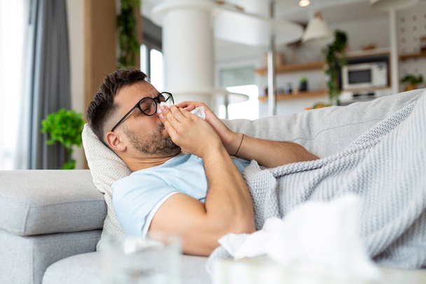 Sick sad man with glasses sits on couch at home suffers from runny nose flu disease coronavirus pandemic covid epidemic sneezes. Unwell guy feeling bad fever virus illness symptoms indoor - Photo, Image