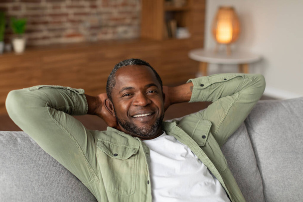 Iloinen keski-ikäinen afrikkalainen amerikkalainen mies lepää sohvalla, nauttia vapaa-aikaa yksin, hiljaisuus ja mukavuus viihtyisä olohuone sisustus, lähikuva, kopioi tilaa. Rentoudu, viikonloppu tauko kotona ja elämäntapa - Valokuva, kuva