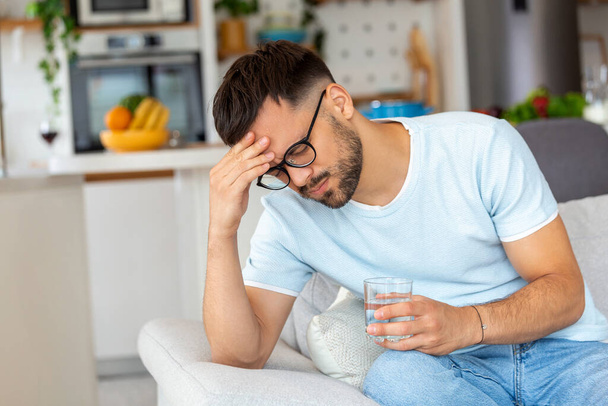 Depressziós fiatalember, akinek fejfájása van, miközben otthon vizet iszik a kanapén. - Fotó, kép