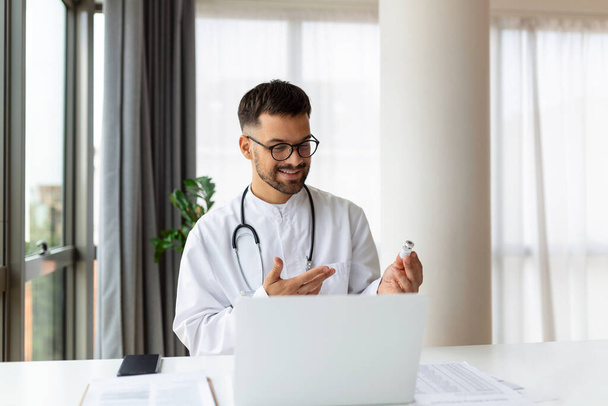 Vriendelijke jonge arts maakt gebruik van laptop om op afstand te communiceren met de patiënt, man in kliniek raadpleegt online, kijkt naar monitor scherm en webcam - Foto, afbeelding