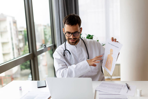 Doctor talking online with patient, showing him digestive system on clipboard, making video call, looking at camera, young male wearing white uniform with stethoscope speaking, consulting and therapy concept - Photo, Image
