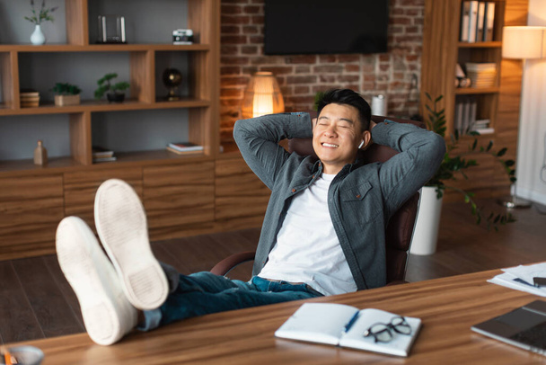 Cheerful confident middle aged asian man in wireless headphones resting at table enjoy free time in home office interior. Relax from business and work, music, audio app during break, covid-19 pandemic - Photo, Image