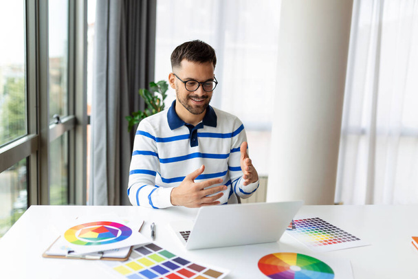 Wesoły projektant mający wideokonferencję z klientami, siedzący przy biurku przed komputerem, trzymający palety kolorów, gestykulujący i uśmiechający się, przestrzeń do kopiowania - Zdjęcie, obraz