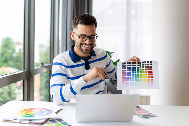 Nuori kaveri osoittaa väri swatches kannettavan tietokoneen näytön kotitoimistossa, esittää graafinen suunnittelu projekti kauko kokouksessa. Millennial suunnittelija valitsemalla gamma tulevaisuuden talon sisustus - Valokuva, kuva